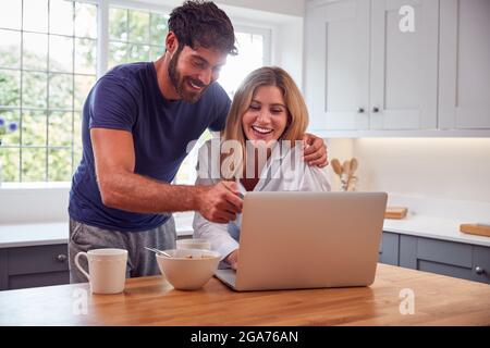 Couple Wearing Pyjamas In Kitchen Watching Laptop Whilst Eating Breakfast Stock Photo