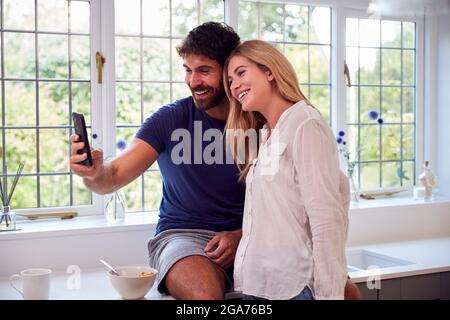 Couple Wearing Pyjamas In Kitchen Using Mobile Phone Whilst Eating Breakfast Stock Photo