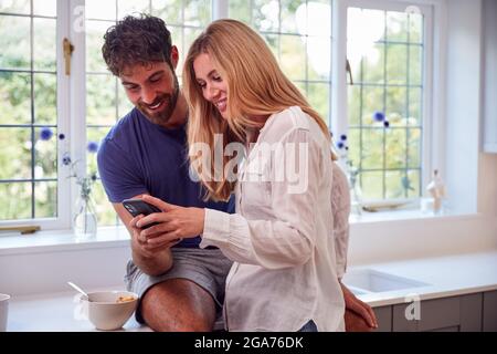 Couple Wearing Pyjamas In Kitchen Using Mobile Phone Whilst Eating Breakfast Stock Photo