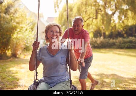 Retired Couple Having Fun With Man Pushing Woman On Garden Swing Stock Photo