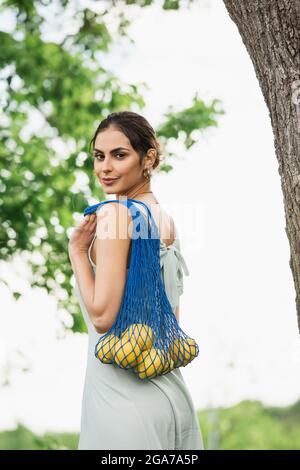 brunette woman holding reusable string bag with lemons in park Stock Photo
