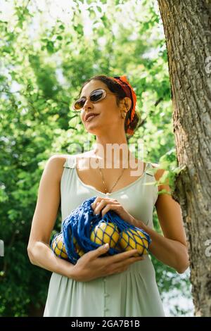 happy woman in sunglasses and dress holding string bag with lemons Stock Photo