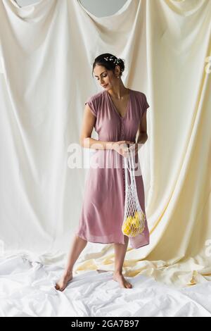 full length of brunette woman holding reusable string bag with lemons on white Stock Photo