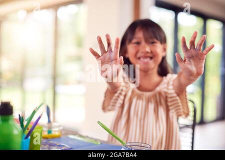 Asian Girl Painting Picture On Table At Home Showing Messy Hands To Camera Stock Photo
