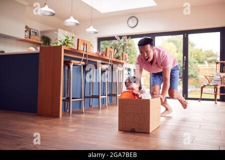 Asian Father And Son Pushing Son Around Kitchen Floor At Home In Junk Modelled Car Stock Photo