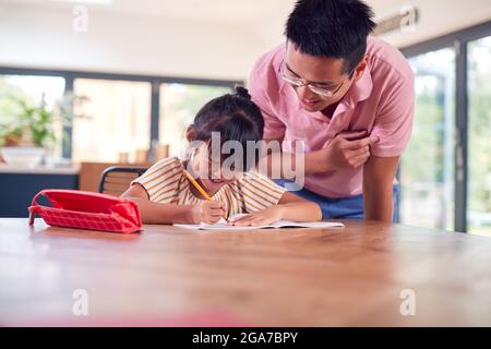 Asian Father Helping Home Schooling Daughter Working At Table In Kitchen Writing In Book Stock Photo