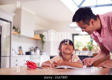 Asian Father Helping Home Schooling Daughter Working At Table In Kitchen Writing In Book Stock Photo