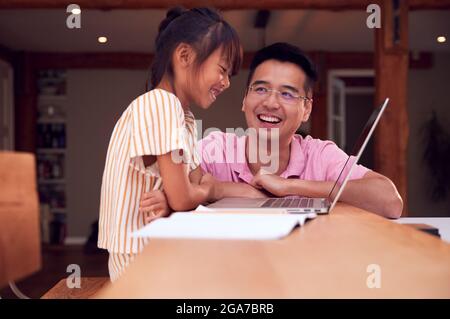 Asian Father Helping Home Schooling Daughter Working At Table In Kitchen On Laptop Stock Photo