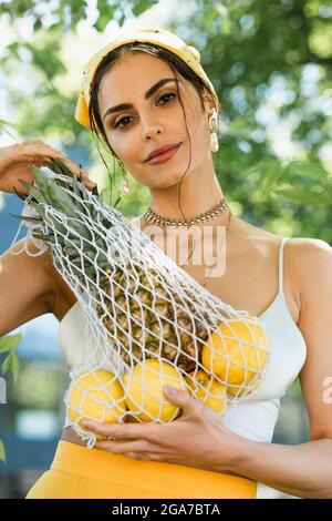 trendy woman in yellow headscarf holding string bag with fruits Stock Photo