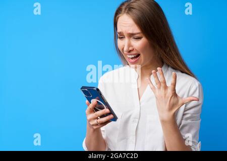 Portrait of young angry woman using her phone, annoyed texting with someone, blue background Stock Photo