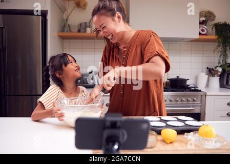 Asian Mother And Daughter Baking Cupcakes In Kitchen At Home Whilst On Vlogging On Mobile Phone Stock Photo