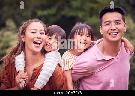 Portrait Of Smiling Asian Family Outdoors With Parents Giving Children Piggybacks Stock Photo