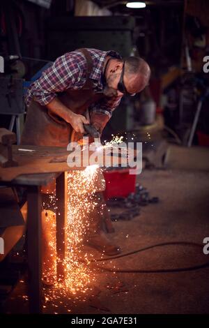 Male Blacksmith Using Plasma Cutter To Cut Shape From Sheet Metal In Forge Stock Photo