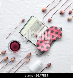 Healthy night sleep. Orange sleep mask with brown polka dots on sleeping time log book. Glass of red herbal tea. Dry poppy flowers, top view on off Stock Photo