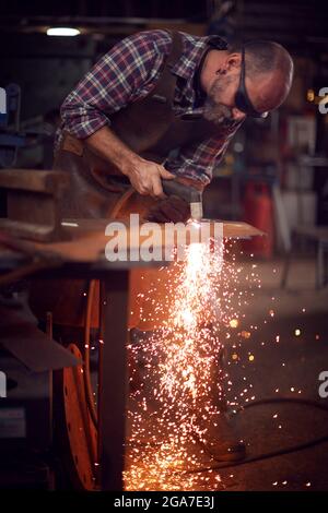 Male Blacksmith Using Plasma Cutter To Cut Shape From Sheet Metal In Forge Stock Photo