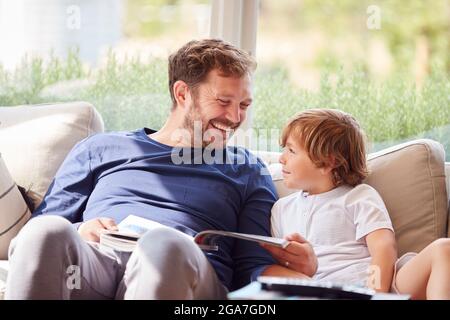 Father And Son Relaxing On Sofa At Home Reading Book Together Stock Photo