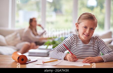 Portrait Of Girl Sitting At Table Home Schooling During Health Pandemic With Mother In Background Stock Photo