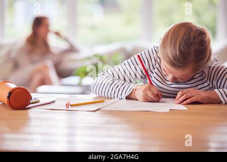 Girl Sitting At Table Home Schooling During Health Pandemic With Mother In Background Stock Photo
