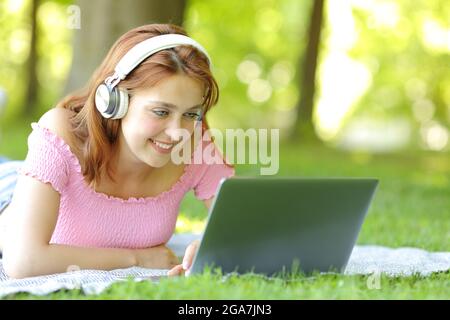 Happy woman wearing wireless headphones checking laptop lying on the grass in a park Stock Photo