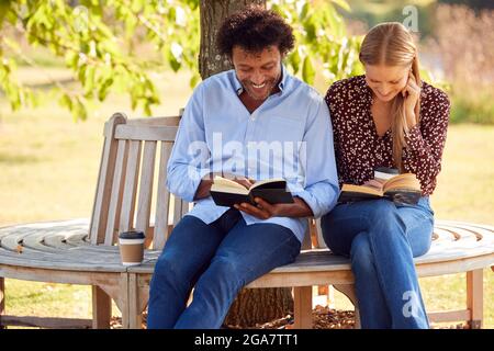 Mature Couple Relaxing Sitting On Park Bench Under Tree Reading Books With Takeaway Coffees Stock Photo