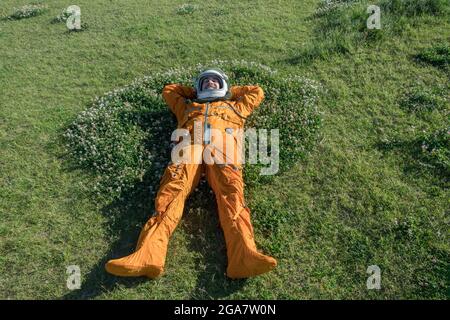 Happy cosmonaut wearing space suit and helmet having a rest while lying on green grass outdoor Stock Photo