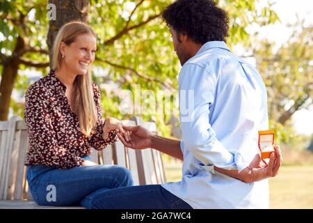 Mature Man Kneeling And Holding Engagement Ring Behind Back About to Propose To Woman In Park Stock Photo