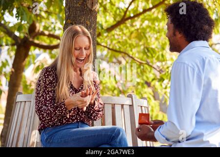 Mature Man Kneeling And Proposing To Surprised Woman Sitting In Park With Engagement Ring In Box Stock Photo