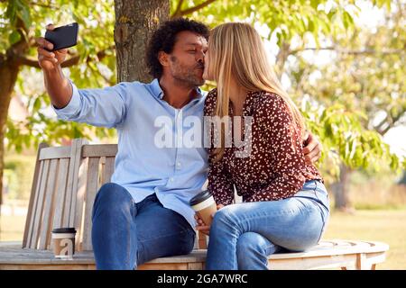 Kissing Mature Couple Posing For Selfie On Mobile Phone Sitting On Seat In Park Stock Photo