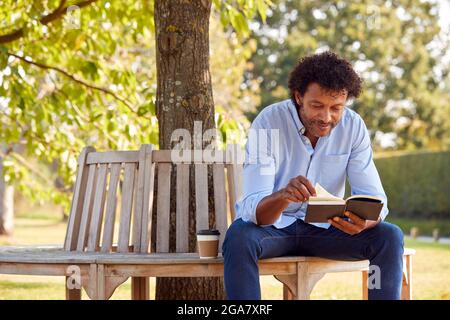 Mature Man Relaxing Sitting On Park Bench Under Tree Reading Book With Takeaway Coffee Stock Photo