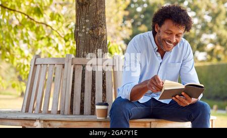 Mature Man Relaxing Sitting On Park Bench Under Tree Reading Book With Takeaway Coffee Stock Photo