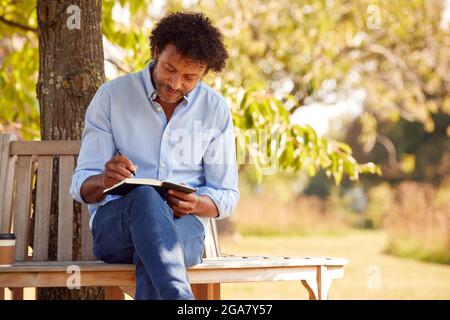 Mature Man Sitting On Park Bench Under Tree Writing In Notebook Stock Photo
