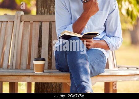 Close Up Of Man With Takeaway Coffee Sitting On Park Bench Under Tree Writing In Notebook Stock Photo