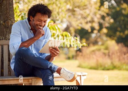 Mature Man Sitting On Bench Under Tree In Summer Park Using Digital Tablet Stock Photo