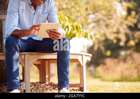 Close Up Of Man Sitting On Bench Under Tree In Summer Park Using Digital Tablet Stock Photo