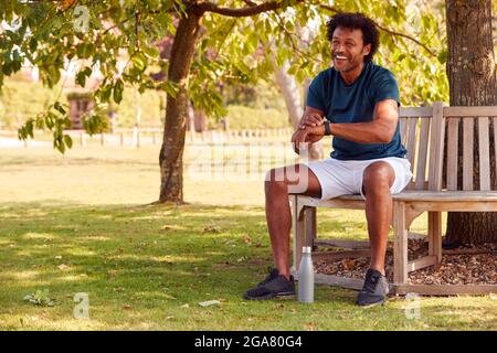 Man Wearing Fitness Clothing Sitting On Seat Under Tree Checking Activity Monitor On Smartwatch Stock Photo