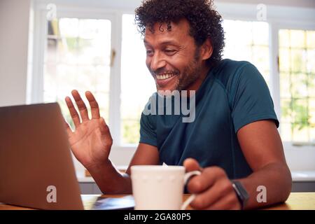 Mature Man At Home In Kitchen Waving As He Makes Video Call On Laptop Computer Stock Photo