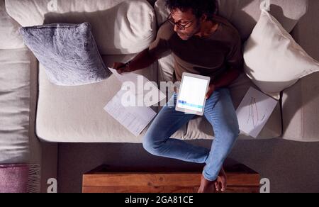 Overhead Shot Of Man Working From Home Sitting On Sofa Reviewing Documents Using Digital Tablet Stock Photo