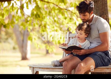 Father Sitting On Park Bench Under Tree With Son Reading Book Together Stock Photo