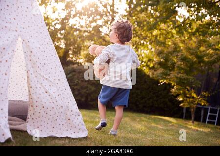 Young Boy Carrying Teddy Bear Having Fun With Tent Or Tepee Pitched In Garden Stock Photo