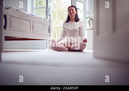 Mature Asian Woman In Pyjamas Sitting On Bedroom Floor Meditating In Yoga Pose Stock Photo