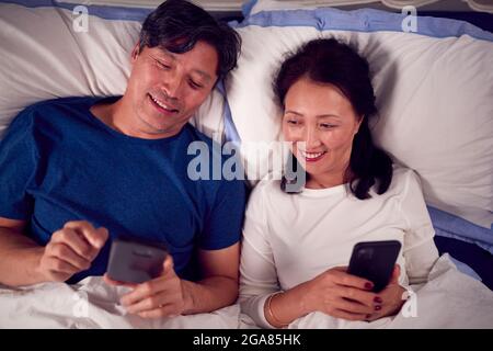 Mature Asian Couple In Wearing Pyjamas Lying In Bed Looking At Mobile Phones Together Stock Photo