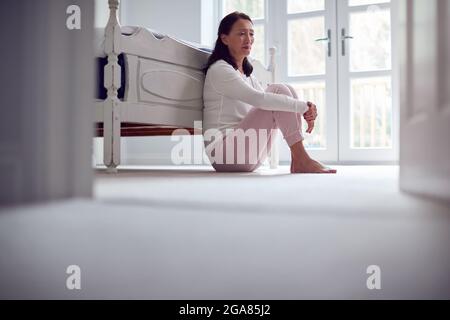 Unhappy And Depressed Mature Asian Woman In Pyjamas Sitting On Bedroom Floor Stock Photo