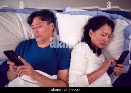 Mature Couple In Wearing Pyjamas Addicted To Using Mobile Phones Lying In Bed And Not Communicating Stock Photo