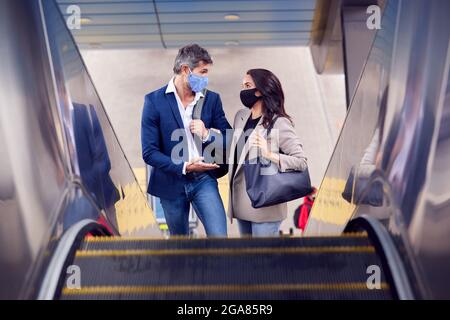 Business Couple Commuting Riding Escalator At Railway Station Wearing PPE Face Masks In Pandemic Stock Photo