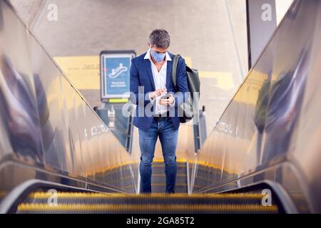 Businessman On Escalator At Railway Station With Mobile Phone Wearing PPE Face Mask During Pandemic Stock Photo