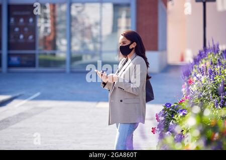 Businesswoman Wearing Mask Walking To Office Looking At Mobile Phone In Health Pandemic Stock Photo