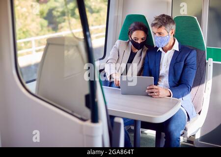 Businesswoman And Businessman On Train Using Digital Tablet Wearing PPE Face Mask During Pandemic Stock Photo