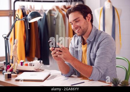 Male Owner Of Fashion Business Using Mobile Phone At Sewing Machine In Studio Stock Photo