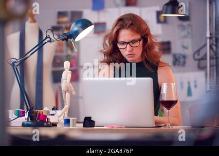 Female Owner Of Fashion Business With Glass of Wine Working Late On Laptop In Studio Stock Photo