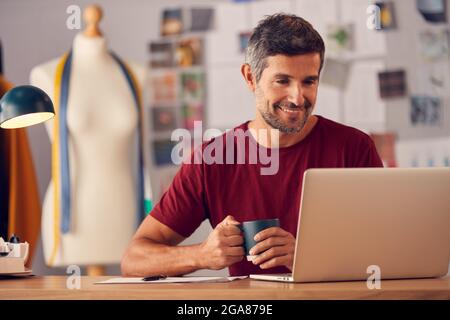 Mature Male Fashion Designer In Studio Sitting At Desk Working On Laptop Stock Photo